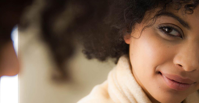 A person with beautiful, curly hair and a neutral expression looks toward the camera, partially visible in a soft-focus background. - Tonya Jones SalonSpa| Birmingham, AL