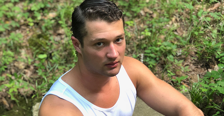 A man with short dark hair, possibly sporting a fresh cut from the salon, is wearing a white tank top and sits outdoors surrounded by green foliage, looking directly at the camera. - Tonya Jones SalonSpa| Birmingham, AL