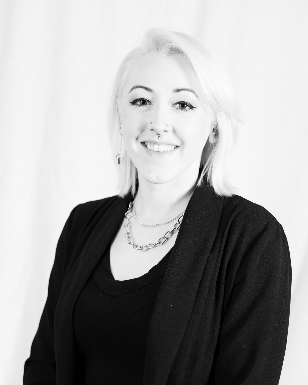 Black and white portrait of a smiling person with short, light-colored hair, a nose piercing, and wearing a dark blouse and jacket. - Tonya Jones SalonSpa| Birmingham, AL