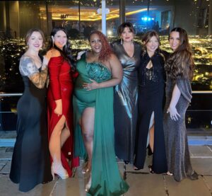 Six women in formal evening gowns pose together indoors, with a cityscape visible through large windows behind them. - Tonya Jones SalonSpa| Birmingham, AL