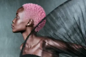 A person with short pink hair and shiny skin poses in profile with a sheer, black, pleated fabric extending behind them against a neutral background. - Tonya Jones SalonSpa| Birmingham, AL
