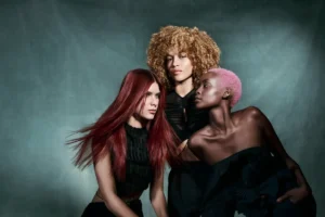 Three women with distinct hairstyles—long red hair, short curly blonde hair, and short pink hair—pose together against a muted green backdrop, wearing black clothing. - Tonya Jones SalonSpa| Birmingham, AL