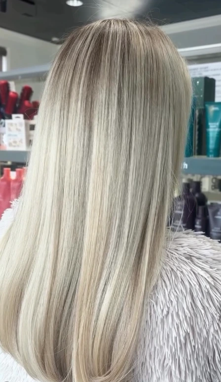 A person with straight, long, light blonde hair is seen from behind, standing in front of shelves with various hair care products. - Tonya Jones SalonSpa| Birmingham, AL