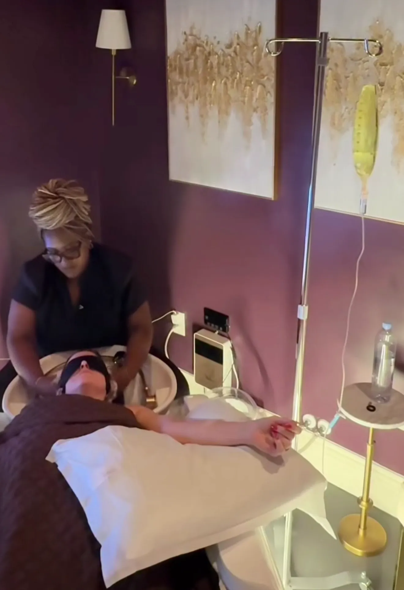 A person lies on a bed receiving a head massage while connected to an IV drip in a room with purple walls and gold-accented artwork. - Tonya Jones SalonSpa| Birmingham, AL